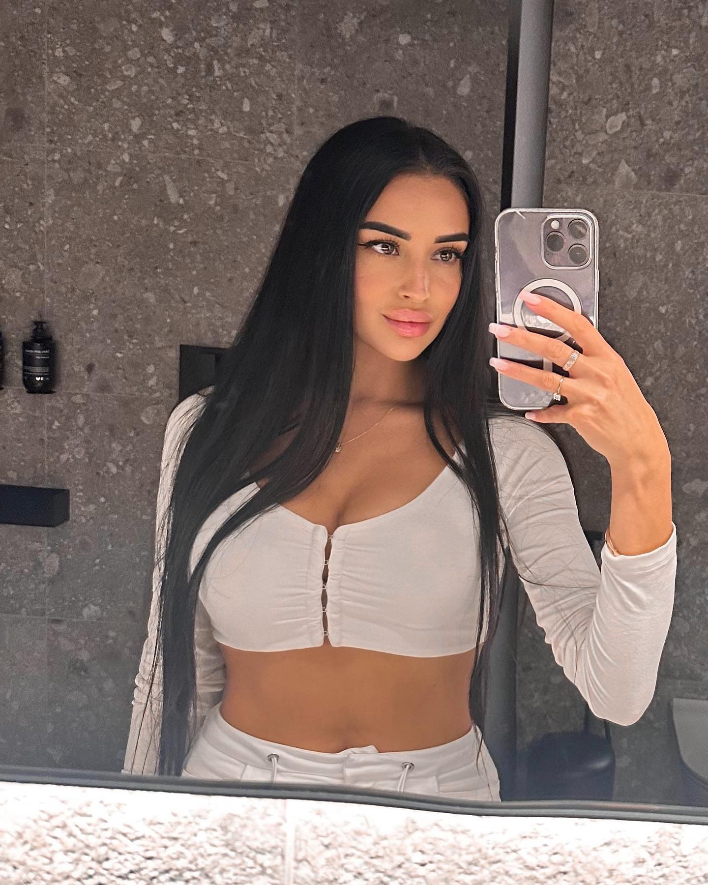 A mirror selfie in a bathroom shows a woman wearing a white cropped button-up top and drawstring pants, holding a smartphone.
