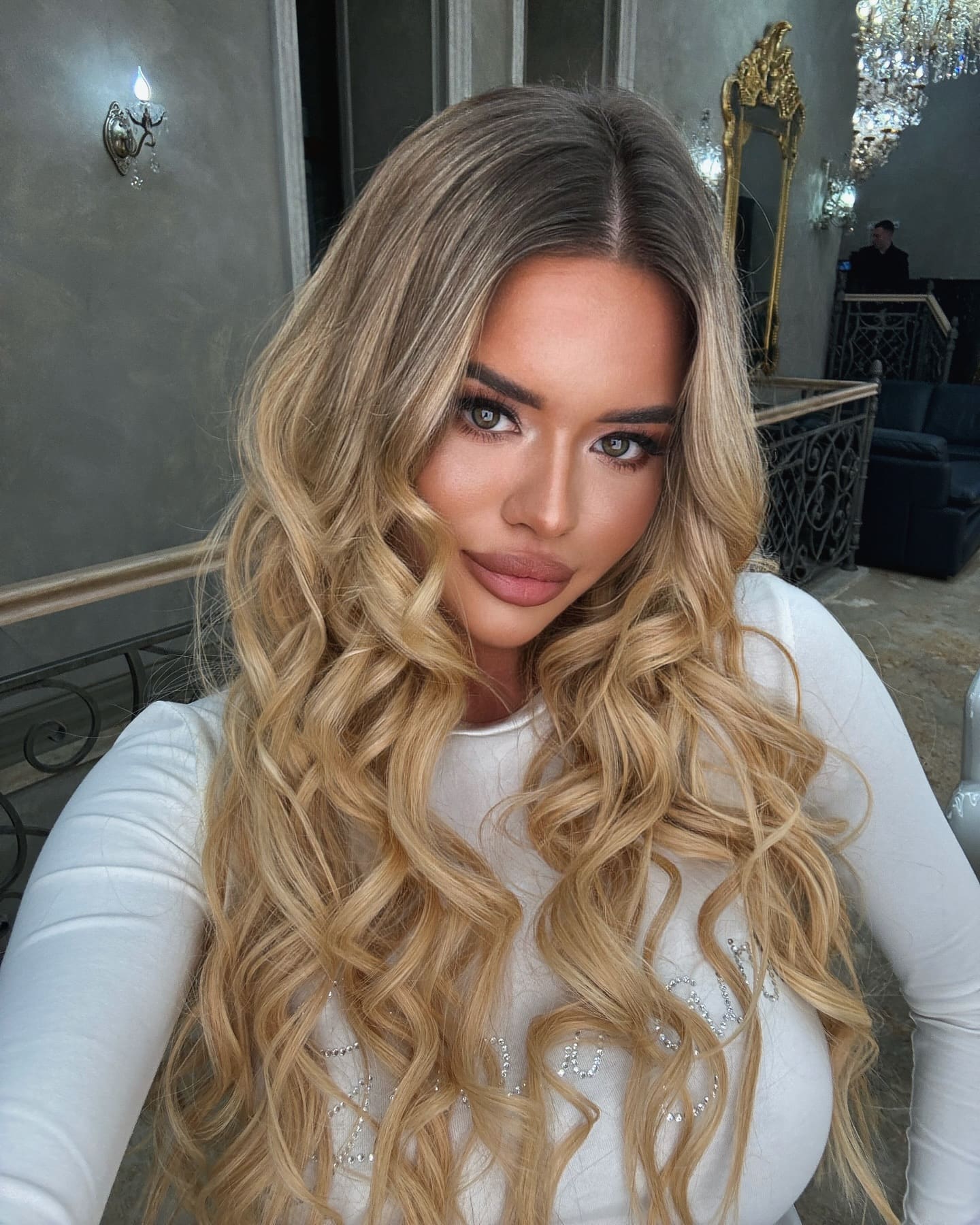 A woman with wavy blonde hair takes a selfie in a white embellished top, standing indoors near a chandelier and gold mirror.