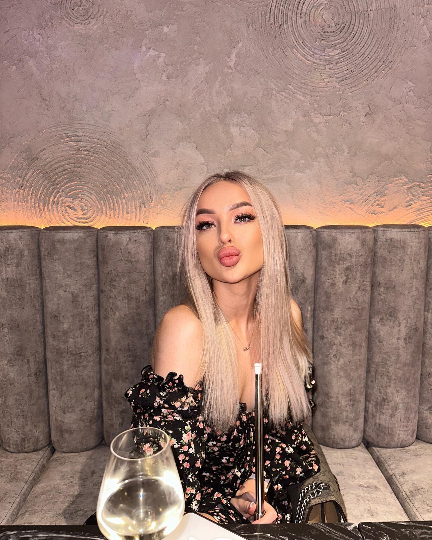 In a restaurant booth, she wears a black off-shoulder floral top, puckering her lips at the camera behind a wine glass and drink.