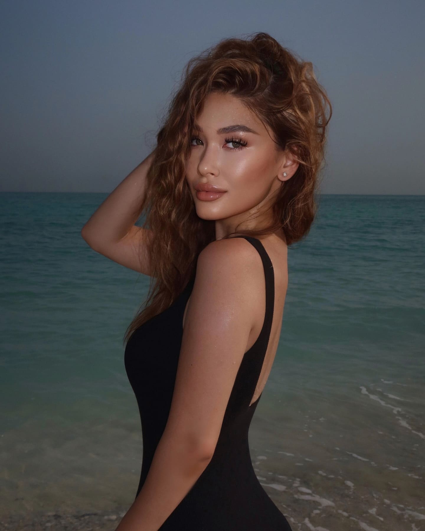 Efia stands in profile at the edge of a calm sea, wearing a sleeveless black one-piece swimsuit with a low back and her right hand raised behind her head.