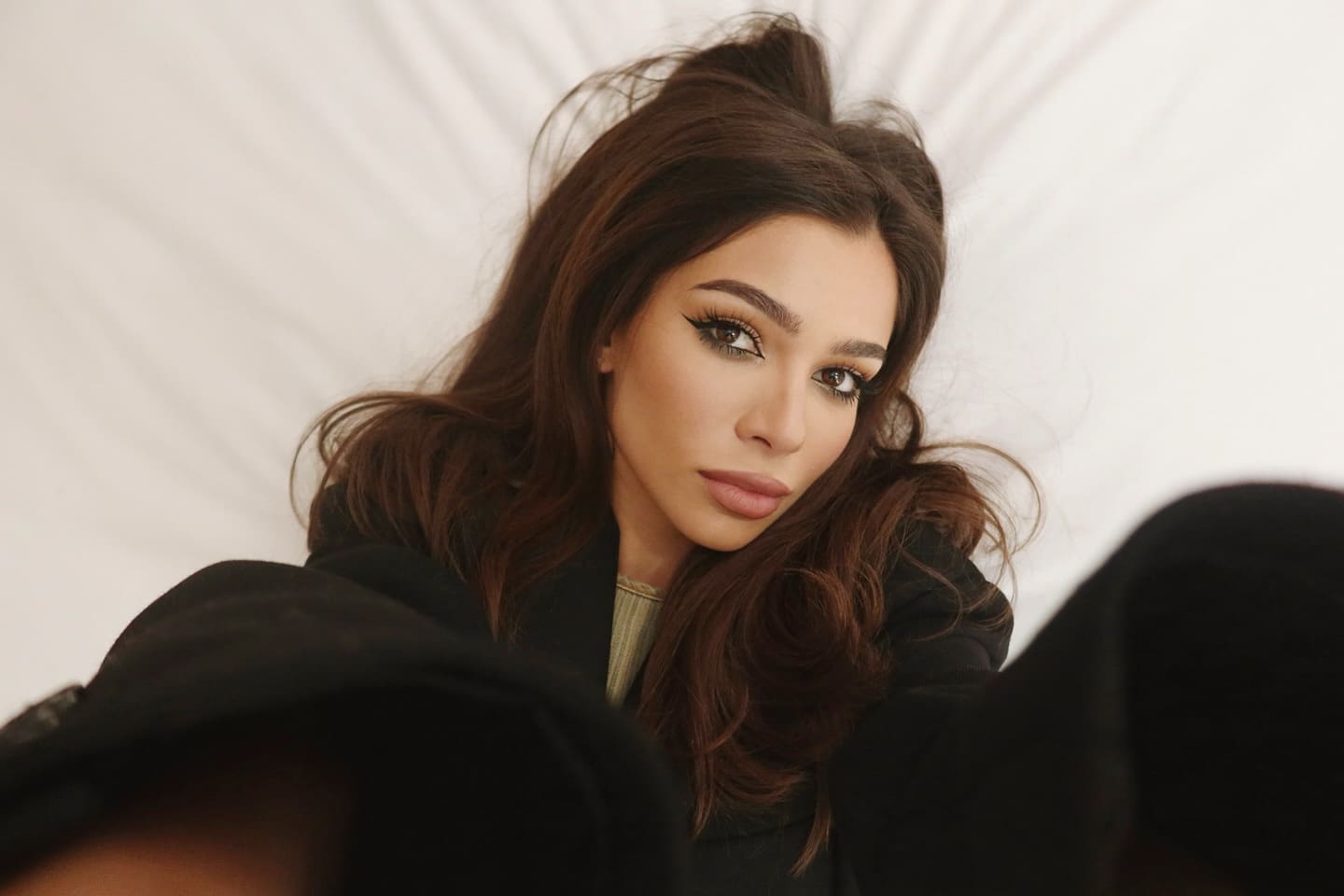Yalya leans forward with crossed arms, wearing a black coat over a light top against a white backdrop.