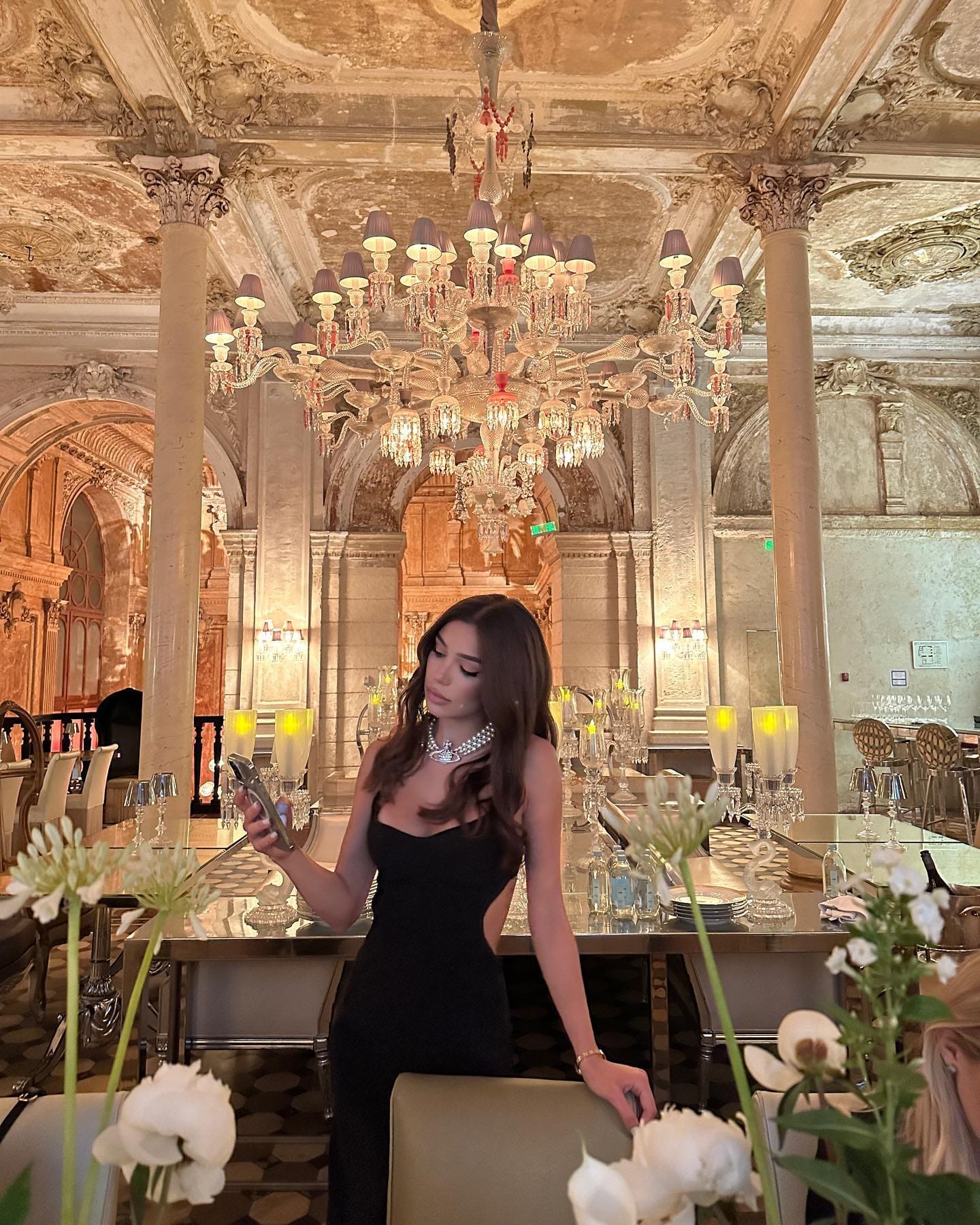 A woman in a sleeveless black dress with a side cutout stands in a candlelit dining hall, holding a phone.