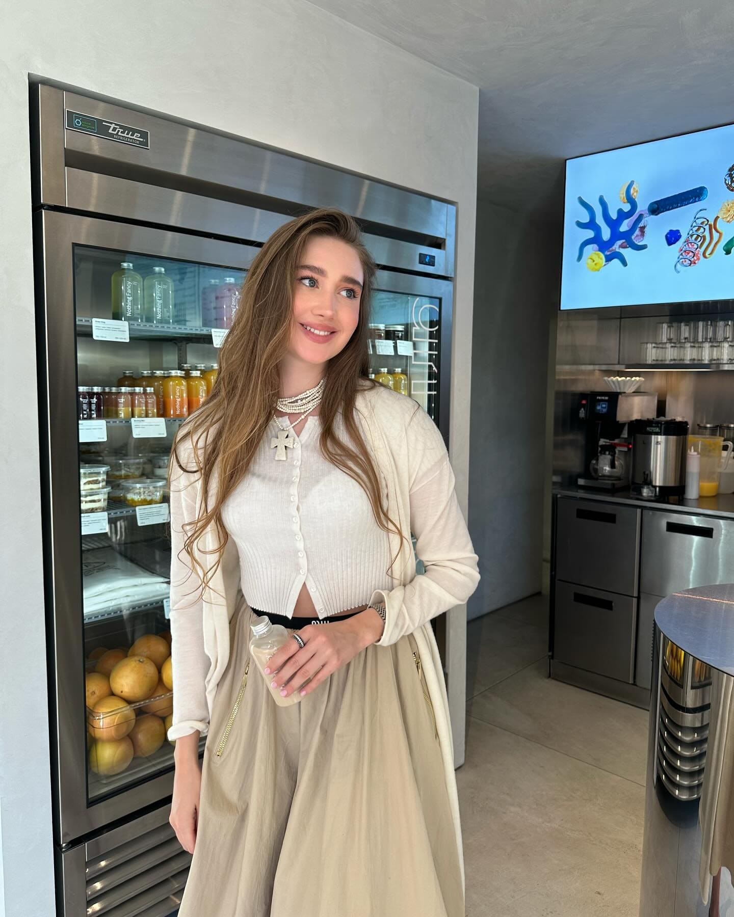 A woman stands in a kitchen holding a small bottle, wearing a white ribbed cardigan, long beige skirt, and pearl necklace.
